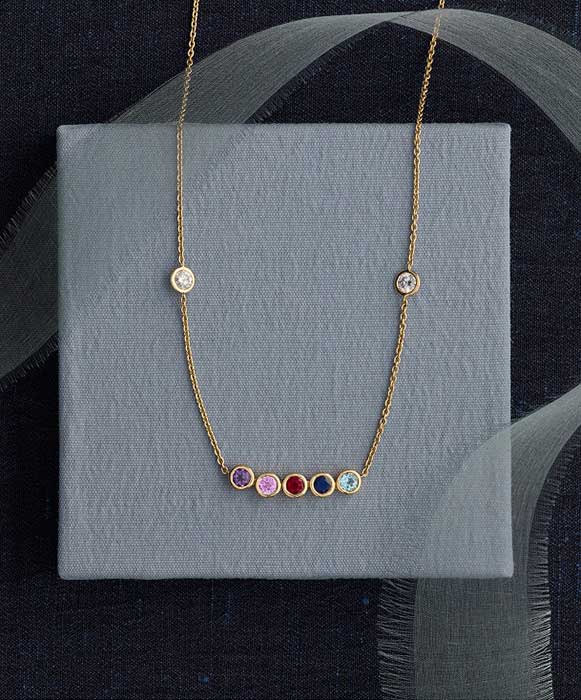 Gold necklace with colorful gemstone charms and diamond accents on a gray display box.